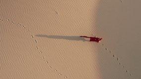 A lonely traveler in fashion red fluttering dress is walking by the beige sand dunes. 4K aerial slow motion video of a beautiful female model in the desert at sunset, California. Nature landscape shot