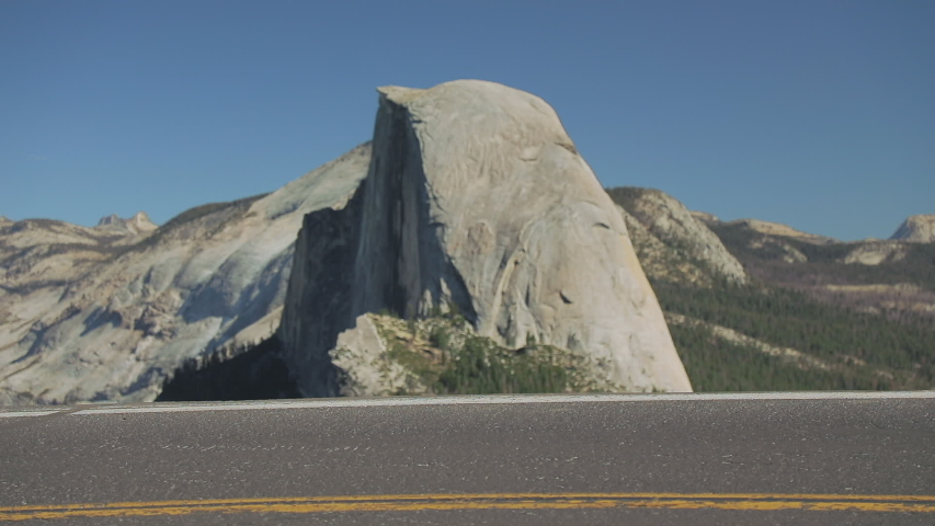 Smiling attractive woman in casual sweatshirt, leggings and jacket is walking through the shot by the edge of road with Half Dome mountain on a background. Slow-motion, Yosemite park California, USA