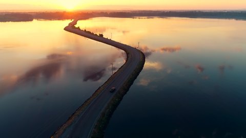 Aerial drone shot of a car driving on a bridge or causeway over a lake at sunrise as the sun peeks over trees on the horizon and clouds reflect in the surface of the water.