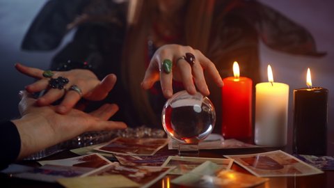 Prediction ritual in dark room with magic crystal ball and candles, gypsy reading hand lines of young man and spelling for happy future, good sorcerer giving her helping hand to helpless man