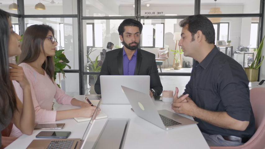 A group of young male and female office employees sitting and discussing on a project in a conference or board room meeting. A team of attractive business partners or people interacting using laptops | Shutterstock HD Video #1056022742