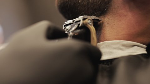 Barber shaving bearded man in barbershop. Male skin care and beard style concept. Master hairdresser with a professional trimmer in a black gloves makes a haircut