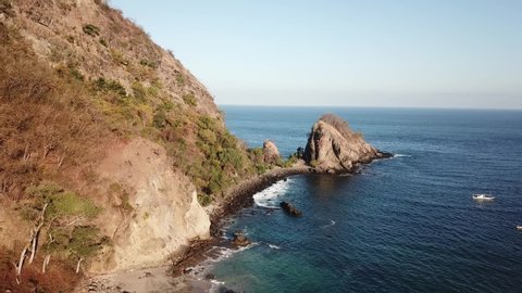 A view on idyllic Koka Beach. Hidden gem of Flores, Indonesia. There is a boat drifting next to the shore. Steep hills going straight into the sea. Few boulders on the open sea. Soft colors of sunset.