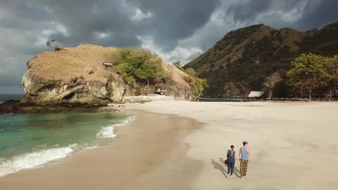 Couple holding hand and walking on idyllic Koka Beach. Hidden gem of Flores, Indonesia. Couple is enjoying their romantic escape. Waves gently washing the shore. Hills in the back. Happiness and love
