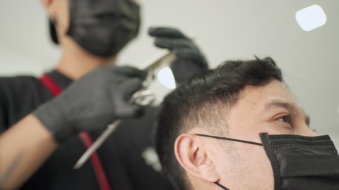 Barber using scissor trimming Asian male customer. close up face guy get a Hair Cut at the Barbershop wear black protective mask. job opportunity, start up business, Pandemic Covid-19 re-open business