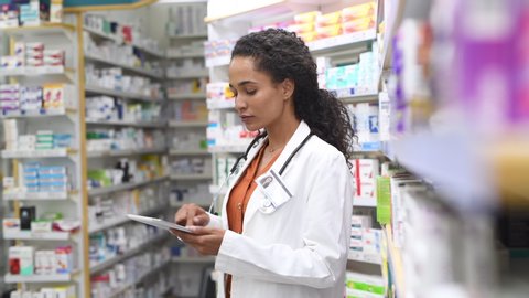 Happy friendly multiethnic pharmacist doing inventory in a provided and modern pharmacy while looking at camera. Portrait african smiling young doctor woman working in drugstore with digital tablet.