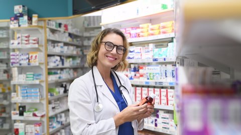 Portrait of mature woman pharmacist at pharmacy wearing labcoat with stethoscope. Happy smiling doctor standing taking medicine from shelf at pharmacy drugstore. Friendly pharmacist looking at camera.
