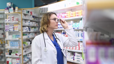 Mature woman pharmacist at pharmacy wearing labcoat with stethoscope. Professional doctor standing taking medicine from shelf at modern pharmacy drugstore.