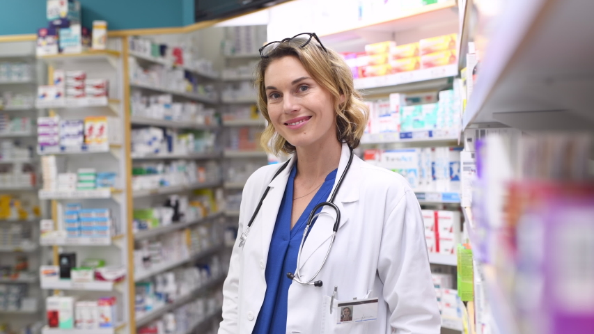 Portrait of mature woman pharmacist at pharmacy wearing labcoat with stethoscope. Happy smiling doctor standing in pharmacy drugstore. Friendly pharmacist owner standing in shop and looking at camera. Royalty-Free Stock Footage #1056031175