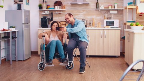 Cheerful disabled woman in wheelchair taking a selfie with husband in kitchen. Hopeful husband with handicapped disabled disability invalid paralysis handicap person next to him, helping her to