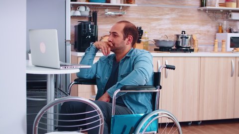 Handicapped businessman in wheelchair using laptop in kitchen. Corporate man with paralysis handicap disability handicapped difficulties working after accident having internet online video call