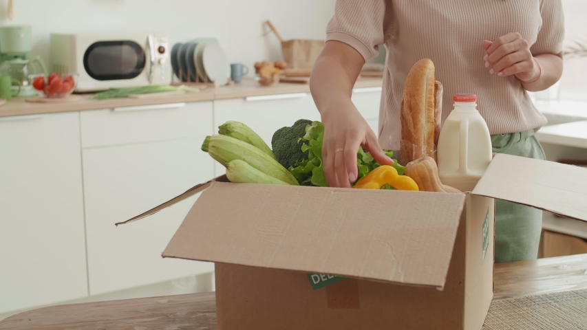 Medium shot of a young mixed-race woman standing at the kitchen table. taking fresh vegetables out of delivered box and putting them on table. | Shutterstock HD Video #1056034628