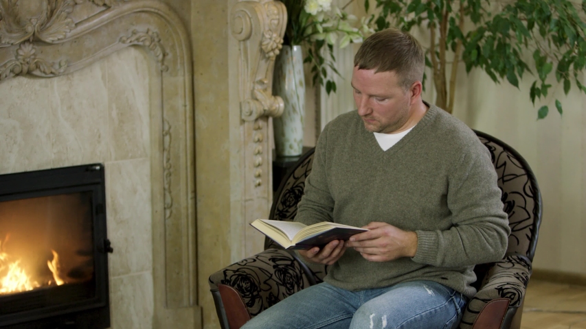 The man sitting in a chair and reading a book in front of the fireplace. Video. Butterfly flying near the male wearing pullover and jeans sitting at home by the fire and reading.