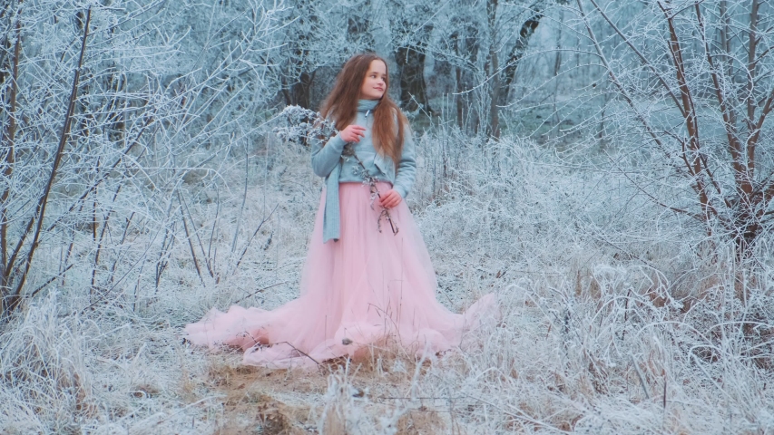 Beautiful charming redhaired cute little girl posing model on backdrop frosty snowy forest. Teenager holding ice tree branch in hands. Warm blue sweater, pink luxury vintage long skirt dress, train