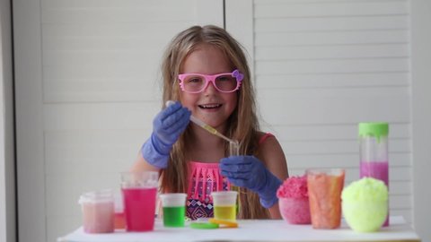 A little girl in pink glasses conducts experiments, looks at the camera and smiles. A child in rubber gloves takes a yellow liquid with a pipette and adds it to the test tube. Experiment.
