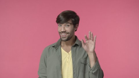 Handsome open-minded, bearded hispanic gay man, friendly guy saying hi shy and silly, smiling greeting person, waving hand hello gesture, introduce himself on new group meeting, pink background