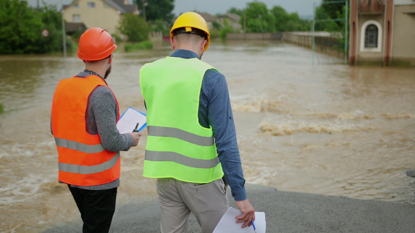 Two male engineers solve flood problem. Devastation wrought after massive disasters. Flooded outskirts of city. Traffic along overflowing with water street. Floods hit town. Heavy rain. Flooding.