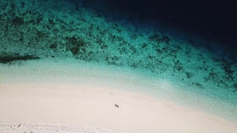 A drone shot of a couple sitting on a pink sand beach in a heart drawn on the sand a small island near Maumere, Indonesia. Happy and careless moments. Waves gently washing the shore. Romance and love
