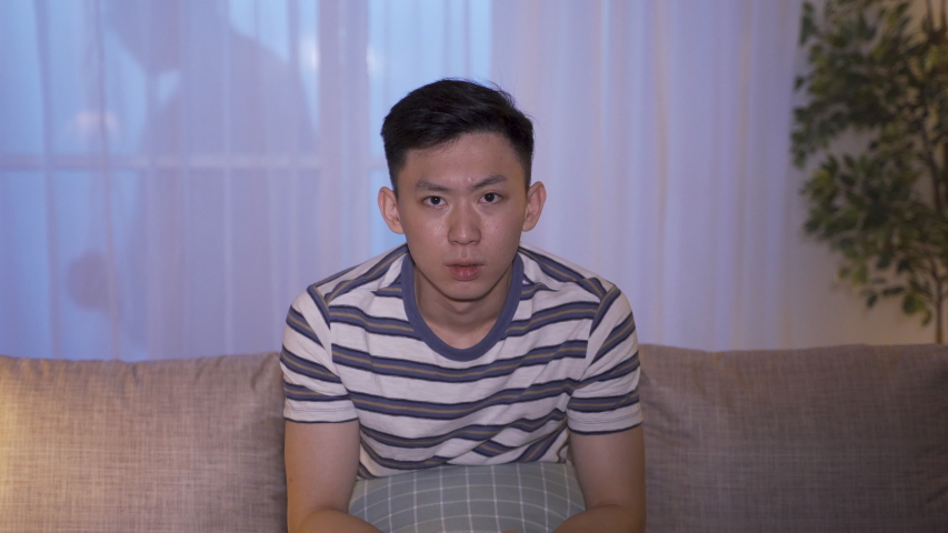 Asian young man got spooky feelings seeing ghost movies alone at nighttime. oddly terrifying atmosphere during horror film suspicious male looking around in house feeling someone is watching him. | Shutterstock HD Video #1056040805