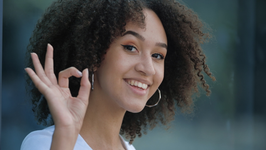 Attractive young afro girl smiles at camera showing hand OK sign. Student shows everything fine success gesture body language. African american millennial curly hair woman smiling, evaluation concept