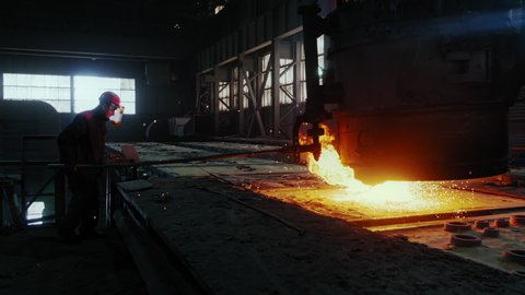 Factory Man in the Foundry of Aluminum Cast, Blacksmith, Bright Yellow Light of Fire and Hot Liquid Molten Metal Pours From Huge Vessel, Sparks, Dark Background, Wide Shot, Slow Motion