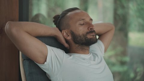 Calm sportive yoga man in white t-shirt sits in a chair in relaxing pose hands behind head close his eyes enjoy peace. Bearded adult guy resting at home after work. Office manager chilling swinging