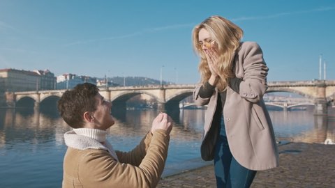 Happy woman taking a proposal about wedding and kissing her future husband. Handsome young man giving an engagement ring to her girlfriend. Couple dating on the bridge. Concept of love and marriage.