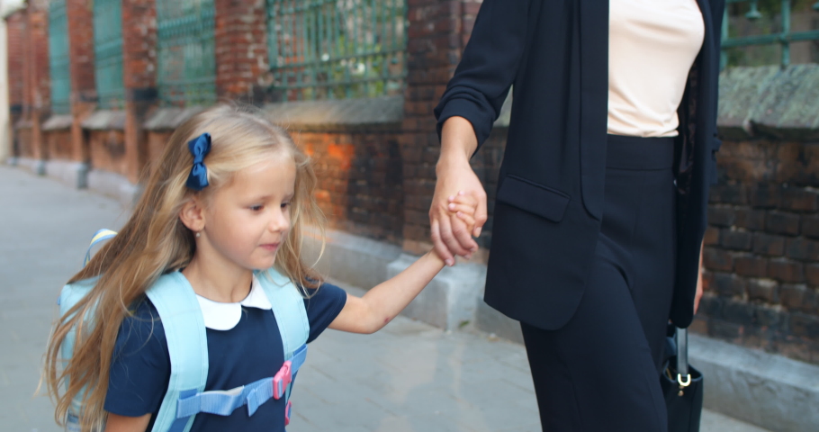 Close up view of pretty child with long blond hair jumping happily and smiling while holding mother hands. Sweet little girl wearing uniform with her mom on their way to school