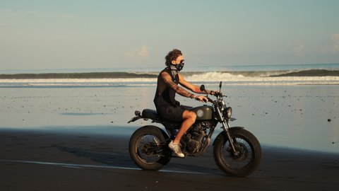 young man is driving motorcycle over sandy beach of ocean, travelling on island