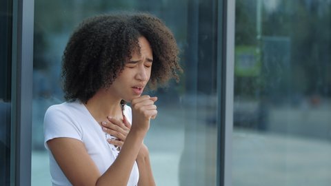 African American girl has sore throat, holds by neck, became ill on street, close up. First symptoms of cold and flu virus, cough appeared. Woman covers mouth face hands, coughs outdoors in city