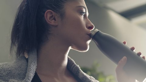 fitness girl drinking from sport bottle during a workout pause at home or gym. Close backlit portrait of active woman drinking from water bottle while resting from training