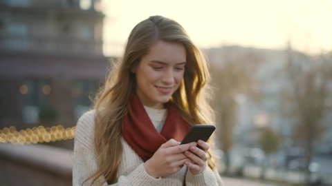 Portrait of happy hipster woman typing by mobile phone outdoors. Closeup cheerful girl walking with smartphone in urban background. Smiling lady holding cellphone in hands outside.