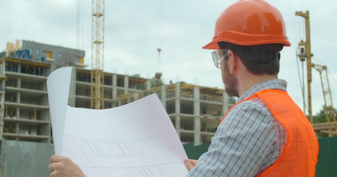 Close-up portrait of engineer with blueprint on background of building under construction. Young architect or builder With Hard Hat Holding Blueprint In His Hands in front of building under