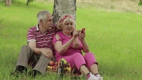 Family weekend picnic. Old people. Senior caucasian couple using smartphone for video call. Talking on webcam. Chatting, social media network on phone. Elderly man, woman. Husband and wife in park