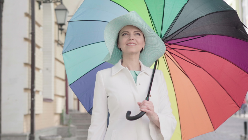 Camera approaches to elegant adult Caucasian woman standing outdoors with colorful umbrella and smiling. Portrait of charming beautiful mid-adult lady enjoying sunny day in city.