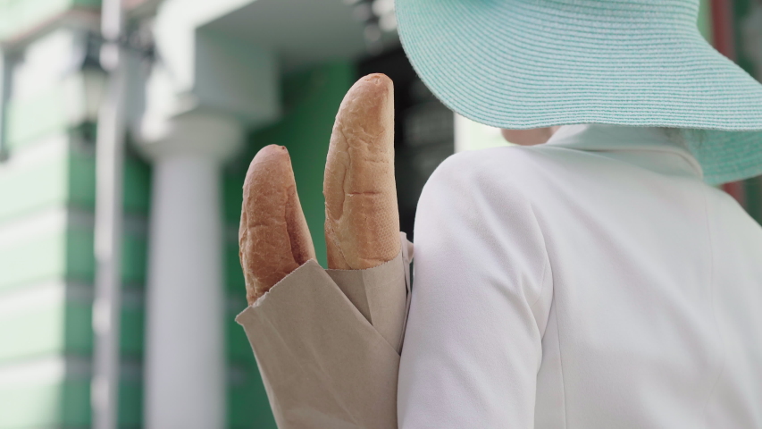 Camera follows unrecognizable elegant lady walking along city street with baguettes. Back side view of happy Caucasian tourist enjoying sunny day outdoors in trip. Leisure, joy, tourism, travelling.