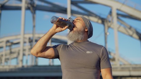 Panning medium shot of senior man with grey beard finishing running workout and drinking water from sports bottle to recover after training