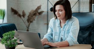 Portrait of Young Attractive Woman looking directly to the Camera while working on her Laptop in modern Loft Office Room. Young Freelance Woman feeling Happy, Satisfied while working.