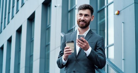Amazing caucasian Businessman is standing near modern Glassy Office Building, holding a Cup of Coffee and using his Smartphone. Successful Businessman in Suit having good Mood, looking satisfied. App.