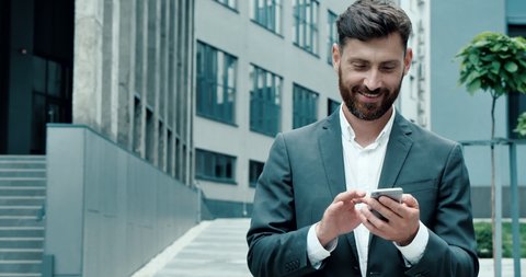 Successful happy Businessman in Luxury grey Suit is texting Messages on his modern Smartphone. Young Man is walking in Business Quartal, having good Mood, looking Satisfied. Successful Lifestyle. Men.