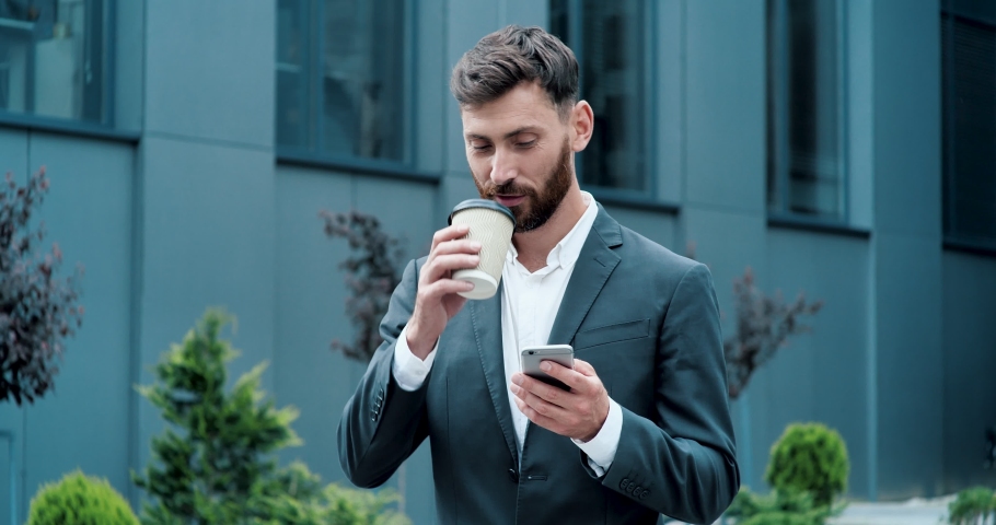 Dark Haired Attractive Business Man with a Beard typing something on his Smartphone, drinking Coffee to go. Concentrated Business Man Reading something in his Phone with cute Smile. Success. Phones. Royalty-Free Stock Footage #1056058487