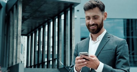 Successful smiled Businessman in Luxury grey Suit is surfing the Internet near the amazing futuristic Office or Business Building. Handsome Young Businessman having Charmingly Smile while using Phone.