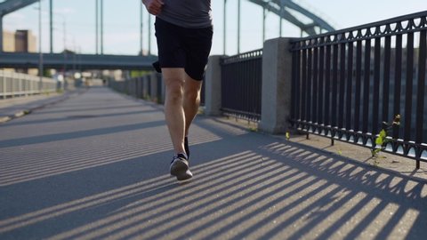 Tilt up slow motion shot of athletic senior man with grey beard running down bridge road in sunlight