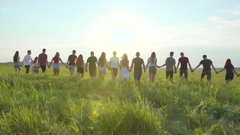 A large group of friends walks through the meadow holding hands in the evening at sunset.