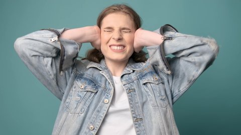Confused woman holding hands on ears over turquoise studio background. Portrait of young serious girl closing her ears, hear no evil, deafness concept.