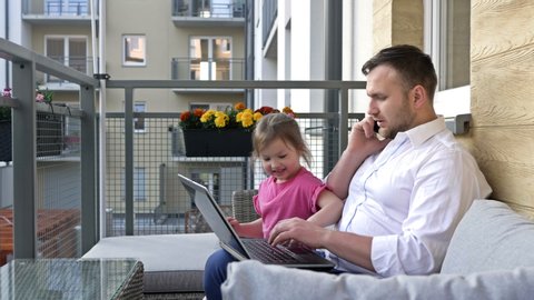 Work remotely. Young man works on the balcony at home. Little daughter wants to play with her dad and prevents him from working.