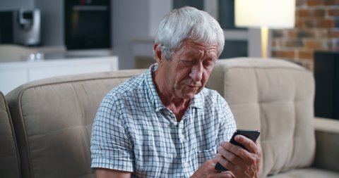 Caucasian old man holds in his hand the smartphone with expression of difficulty in using it. Communication with relatives through video communication. Portrait of a pinseoner man