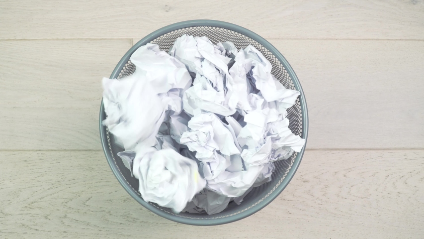 Throw crumpled into the trash. Overflowing waste paper in office garbage bin. Junk, wastepaper in rubbish isolated on wooden background | Shutterstock HD Video #1056066002
