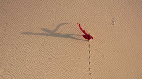 Lonely woman in fashion red fluttering dress is walking by the golden sand dunes. 4K aerial commercial slow motion shot of a beautiful model in the desert at sunset, California. Nature landscape video