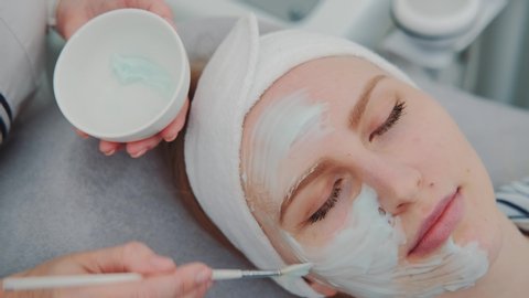Close-up shot of Cosmetician hands applying cream mask on young woman's face at beauty spa salon. Facial skin care treatments.